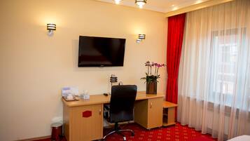 Habitación de hotel con escritorio de madera, televisión de pantalla plana, cortina roja y ventana con cortinas translúcidas.