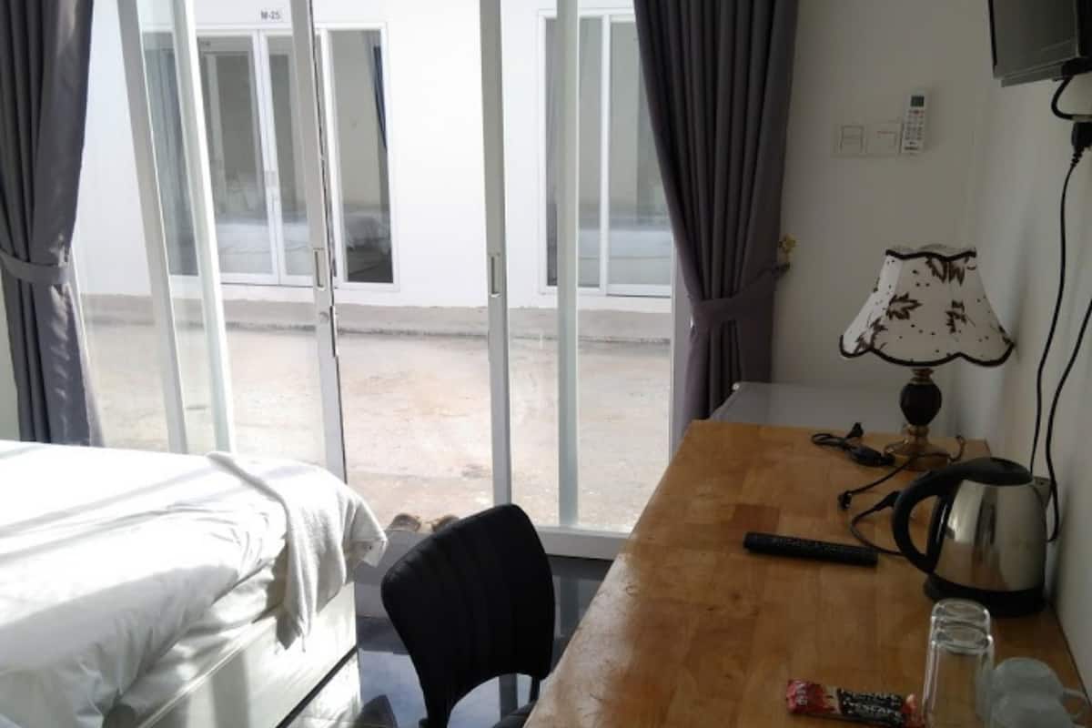 A hotel room with a bed, a wooden desk, a chair, a lamp, a kettle, and a window with curtains.