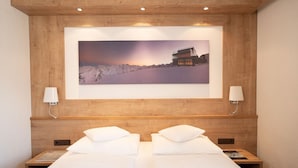 A hotel room with a wooden headboard, a large framed picture of a mountain landscape, two bedside tables with lamps, and a bed with white linens. - Garbershof (Soelden)