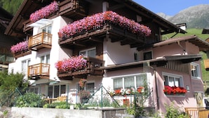 A traditional chalet with multiple balconies adorned with colorful flowers, a stone wall in front, and a mountainous backdrop. - Garbershof (Soelden)