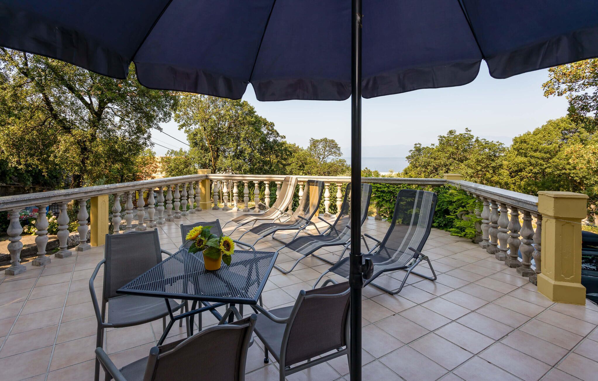 An outdoor patio with a table and chairs, a large umbrella, and a view of trees and a body of water.