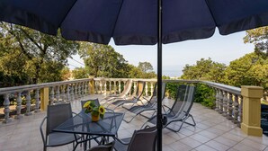 An outdoor patio with a table and chairs, a large umbrella, and a view of trees and a body of water. - Beautiful apartment in Kostrena (Kostrena)