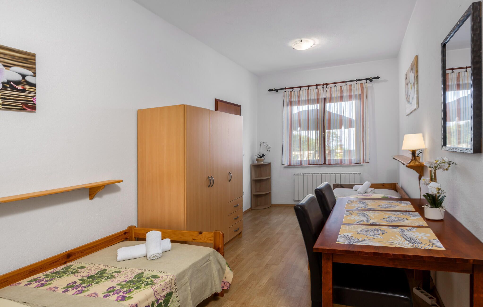 Een kleine, verzorgde kamer met een houten garderobe, een enkel bed met een bloemenbedekker, een houten eettafel met een tafelkleed met dessin, en een raam met gordijnen.