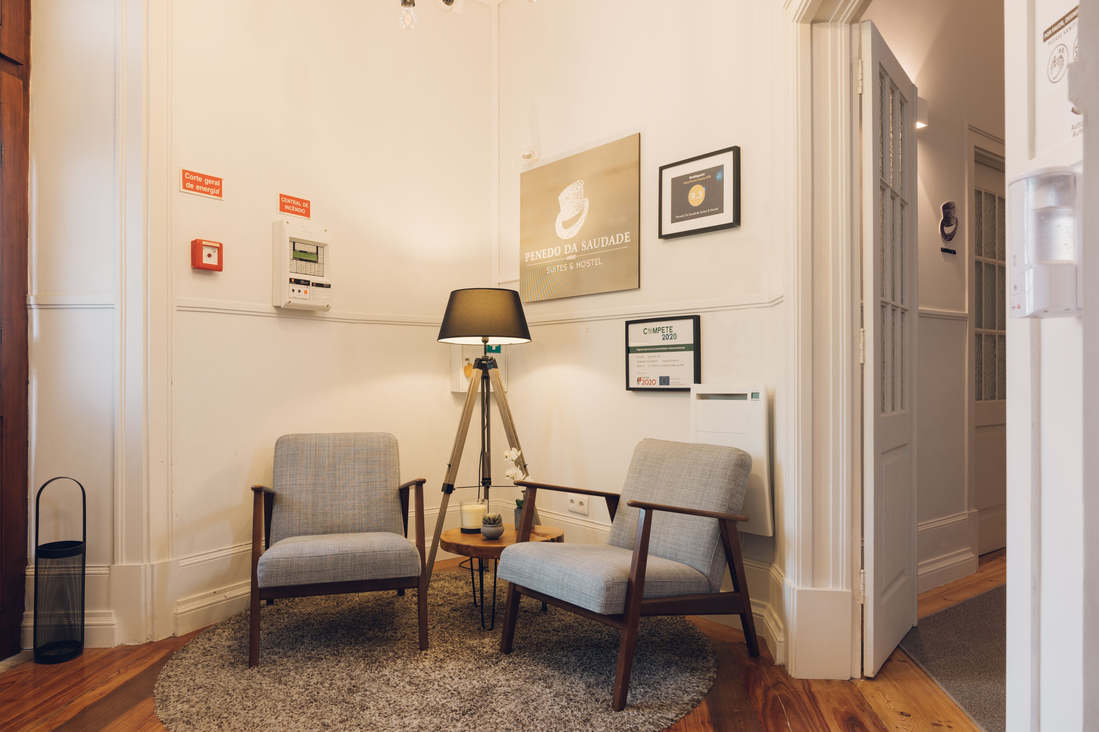 Habitación de hotel con dos sillones grises, una mesita redonda pequeña, una lámpara de pie y una alfombra. Se observan carteles en la pared que indican características de ahorro energético y un certificado enmarcado.