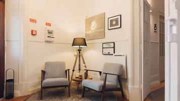 Habitación de hotel con dos sillones grises, una mesita redonda pequeña, una lámpara de pie y una alfombra. Se observan carteles en la pared que indican características de ahorro energético y un certificado enmarcado.