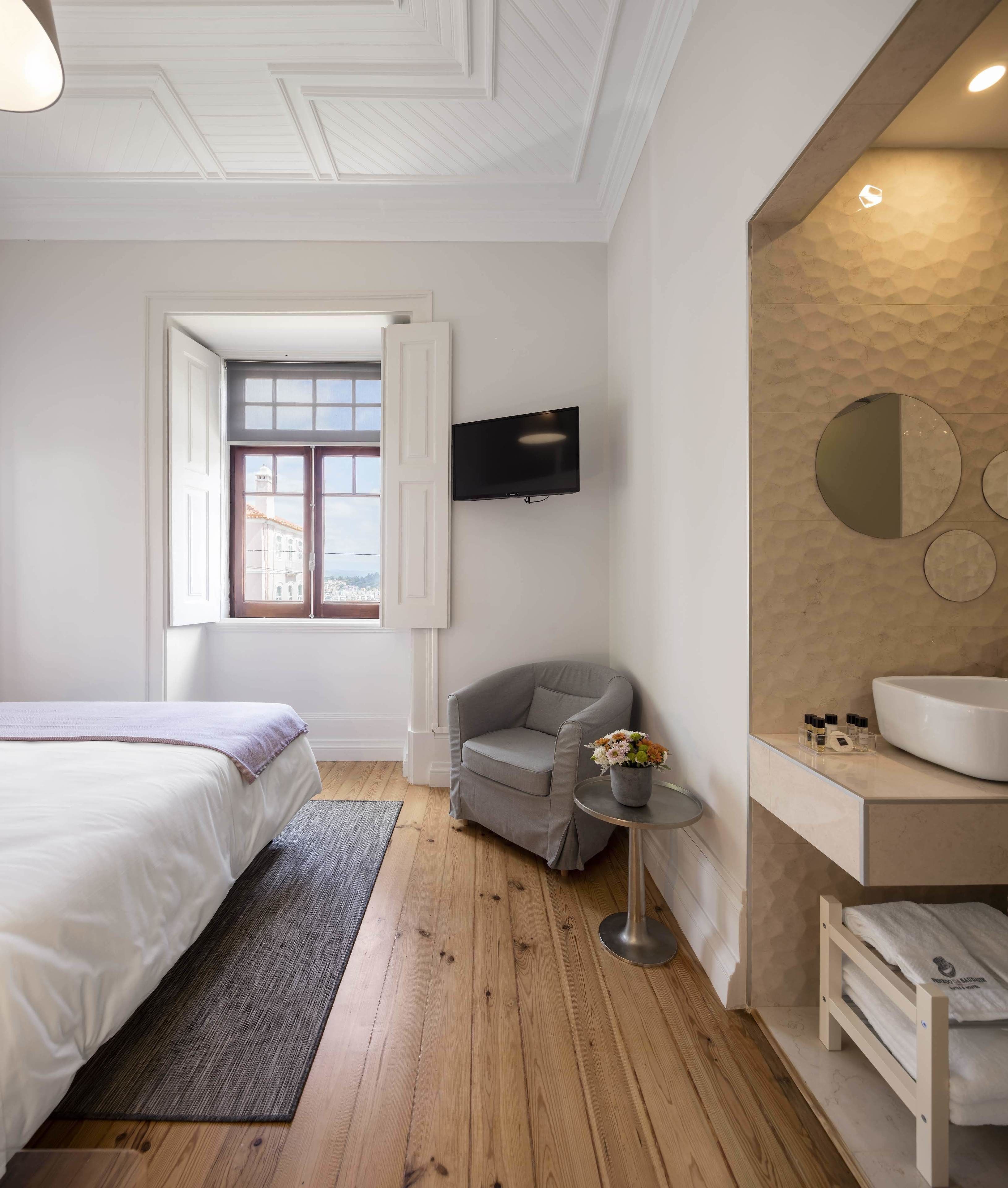 Una habitación de hotel moderna con una cama grande, un sillón gris, una pequeña mesa redonda con un florero, un televisor montado en la pared y un baño con un lavamanos y espejo grandes.