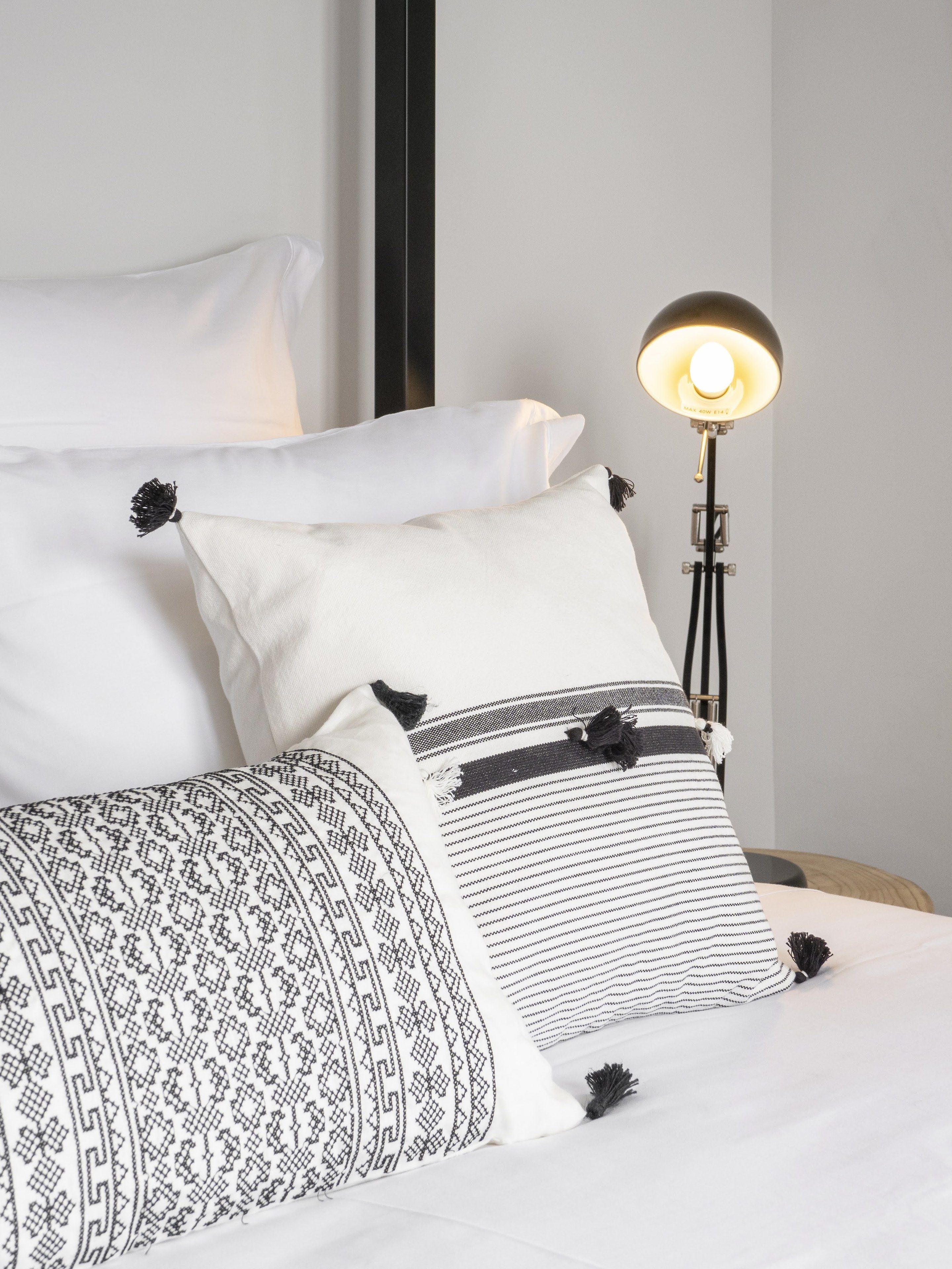 Una cama bien hecha con ropa de cama blanca y estampada, una lámpara negra y una pared lisa.