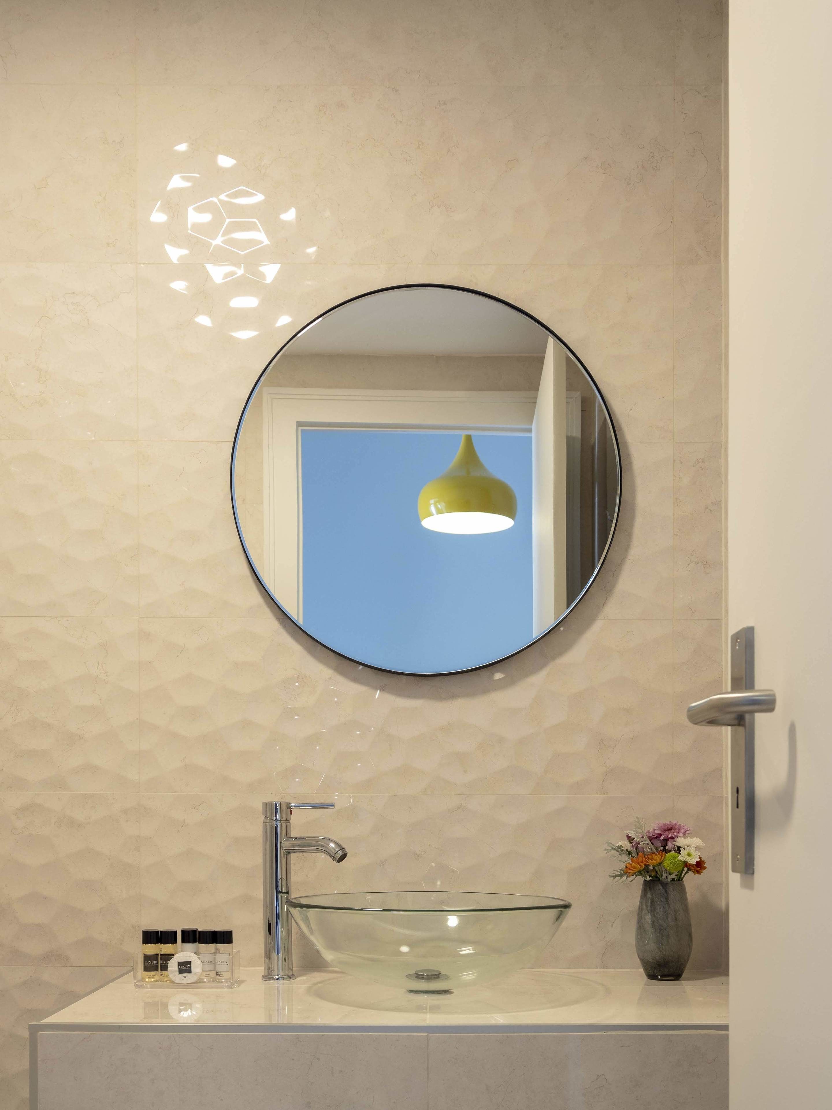 A bathroom with a round mirror, a glass sink, and a vase with flowers.