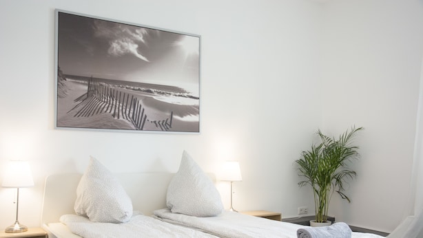 A double bed with white linens and two pillows, a bedside table with a lamp, a wall-mounted picture of a beach, and a potted plant. - Bed'n'Work Apartment Prenzlauer Berg (Berlin)