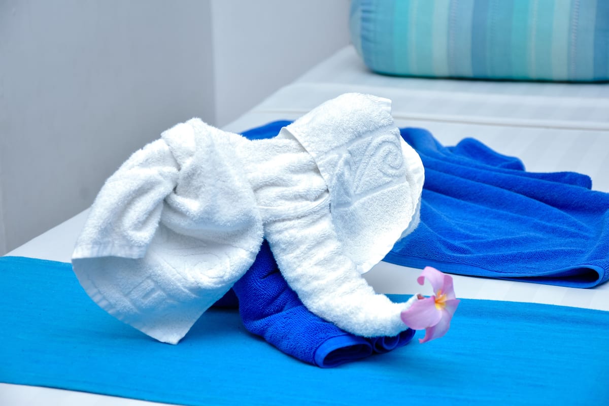 Une serviette blanche et une serviette bleue, pliées avec soin, sont posées sur une surface bleue, qui pourrait être un lit ou une table. Un coussin à rayures se trouve à l’arrière-plan.