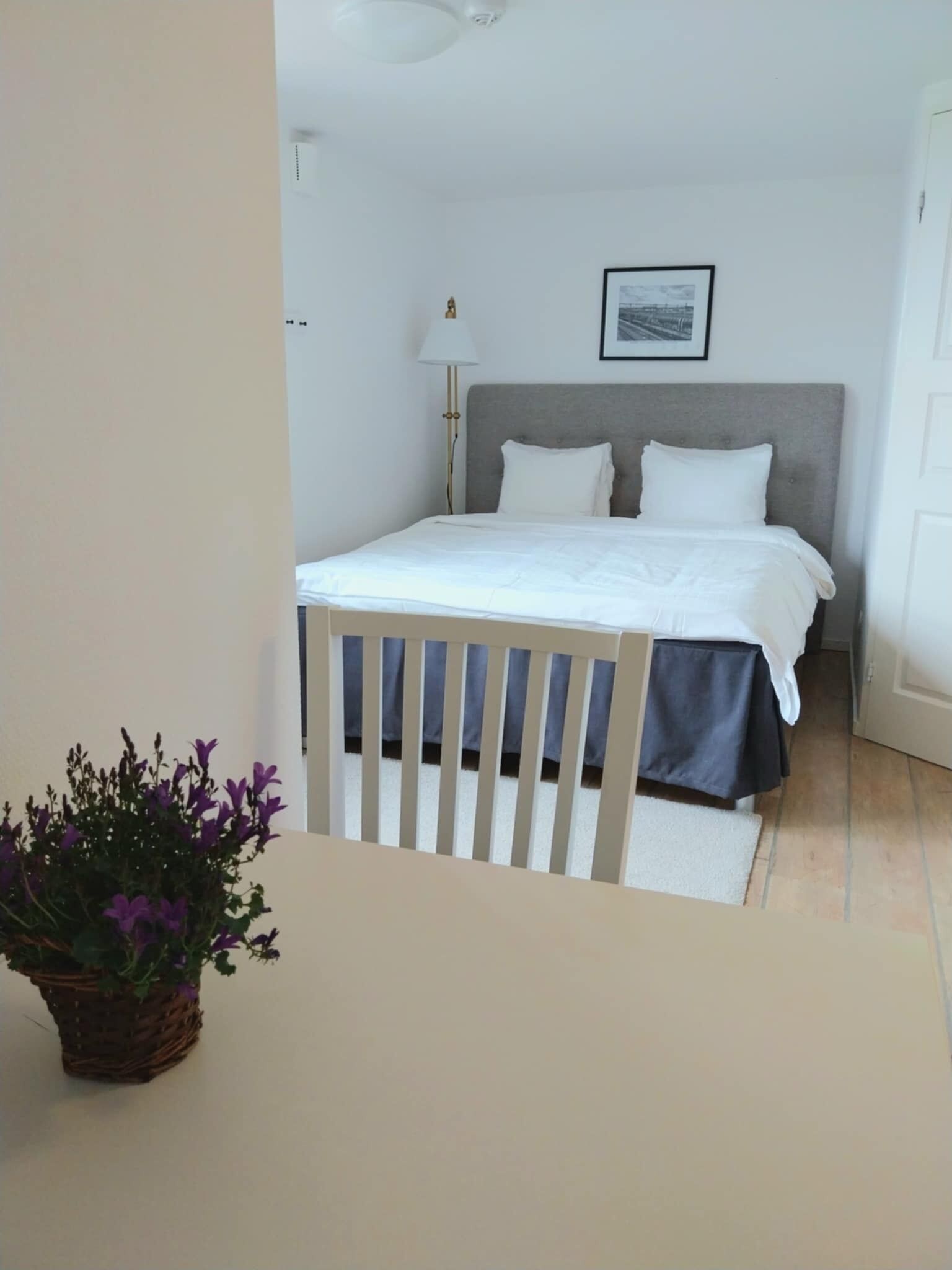 A bedroom with a bed, a bedside lamp, a framed picture on the wall, and a potted plant.