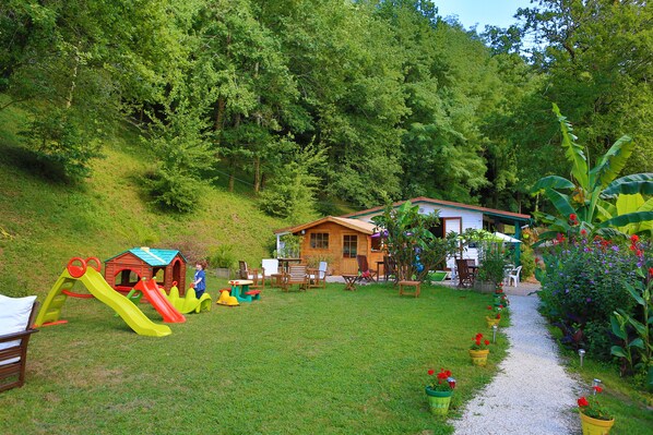 A wooden house with a red roof, surrounded by a grassy yard with a slide and play structures.