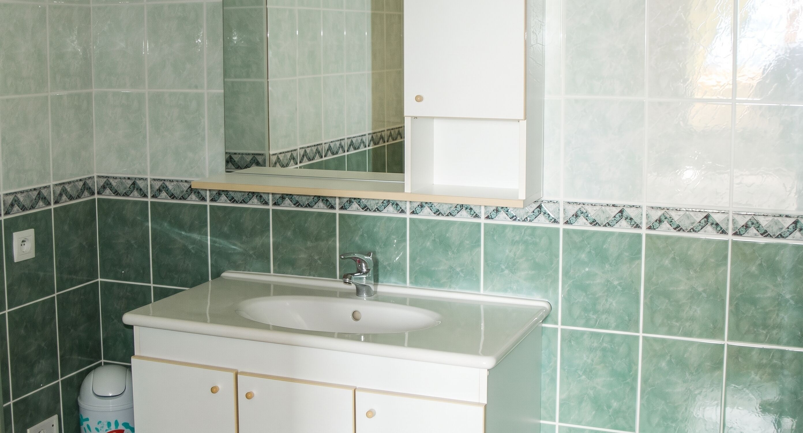 Une salle de bain avec un lavabo blanc, des carreaux verts, un miroir et une poubelle.