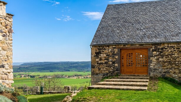A stone building with a wooden door and steps leading up to it, set against a rural landscape with fields and hills.
