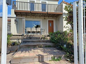 A two-story house with a wooden balcony, a front door, and a small patio with outdoor furniture.