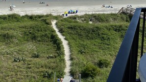 Vista de la playa con un sendero despejado que lleva a la orilla, algunos sombrillas y un velero a lo lejos.