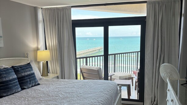 Habitación de hotel con cama, mesita de noche, lámpara, cómoda y vistas al océano desde un balcón.