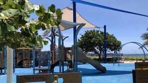 Un parque infantil con tobogán y estructuras para escalar, rodeado de árboles y vistas a la playa.