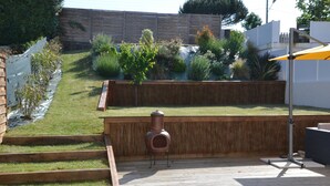 Eine Holzterrasse mit Grill, überdachter Sitzgruppe und Garten mit verschiedenen Pflanzen sowie einem Holzzaun im Hintergrund.