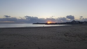 Ein Strand mit Sonnenuntergang im Hintergrund, eine Mole mit Booten und einer Stadtansicht gegenüber dem Wasser.