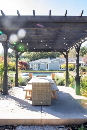 Un patio techado con mesa de billar, mobiliario exterior y vista hacia una casa al fondo.