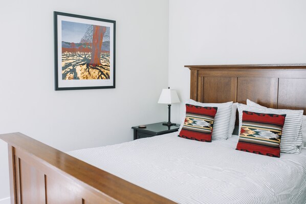 Una cama bien hecha con ropa de cama a rayas y dos almohadones decorativos, un cabecero de madera, un cuadro en la pared y una mesita de noche con una lámpara.