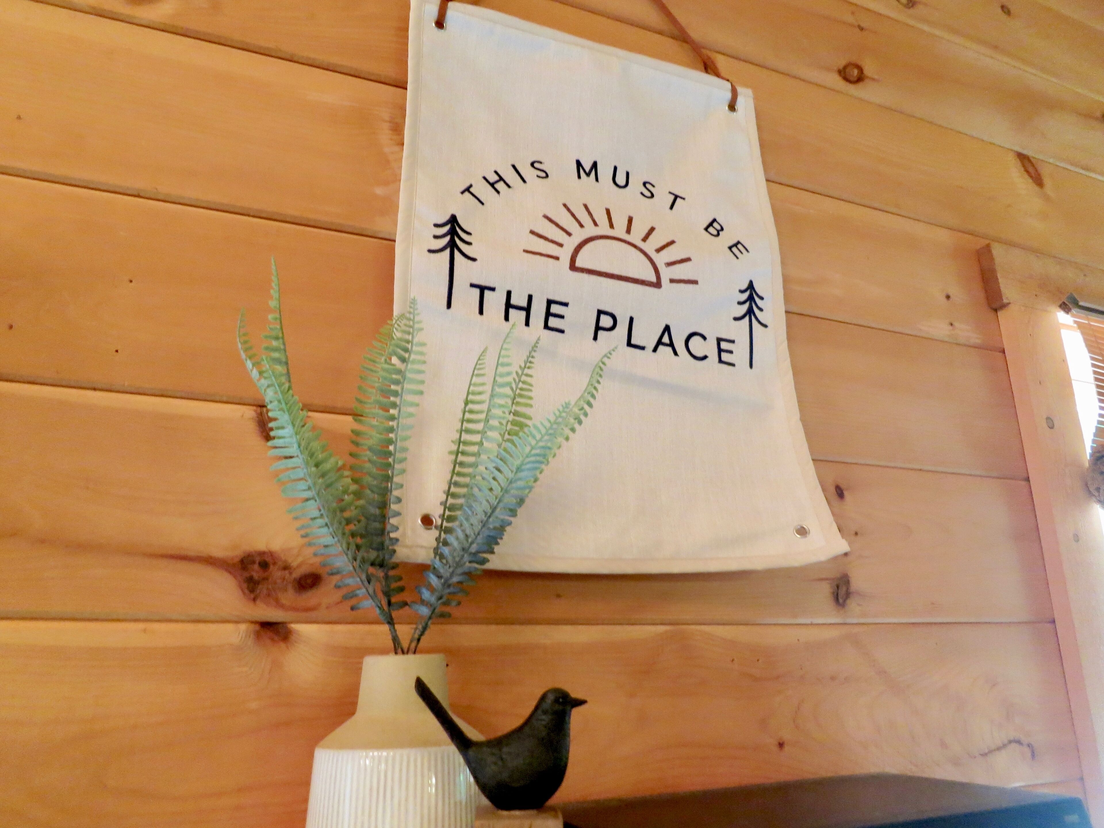 A wooden wall with a hanging sign that reads 'THIS MUST BE THE PLACE' and a white vase with a fern and a bird figurine.