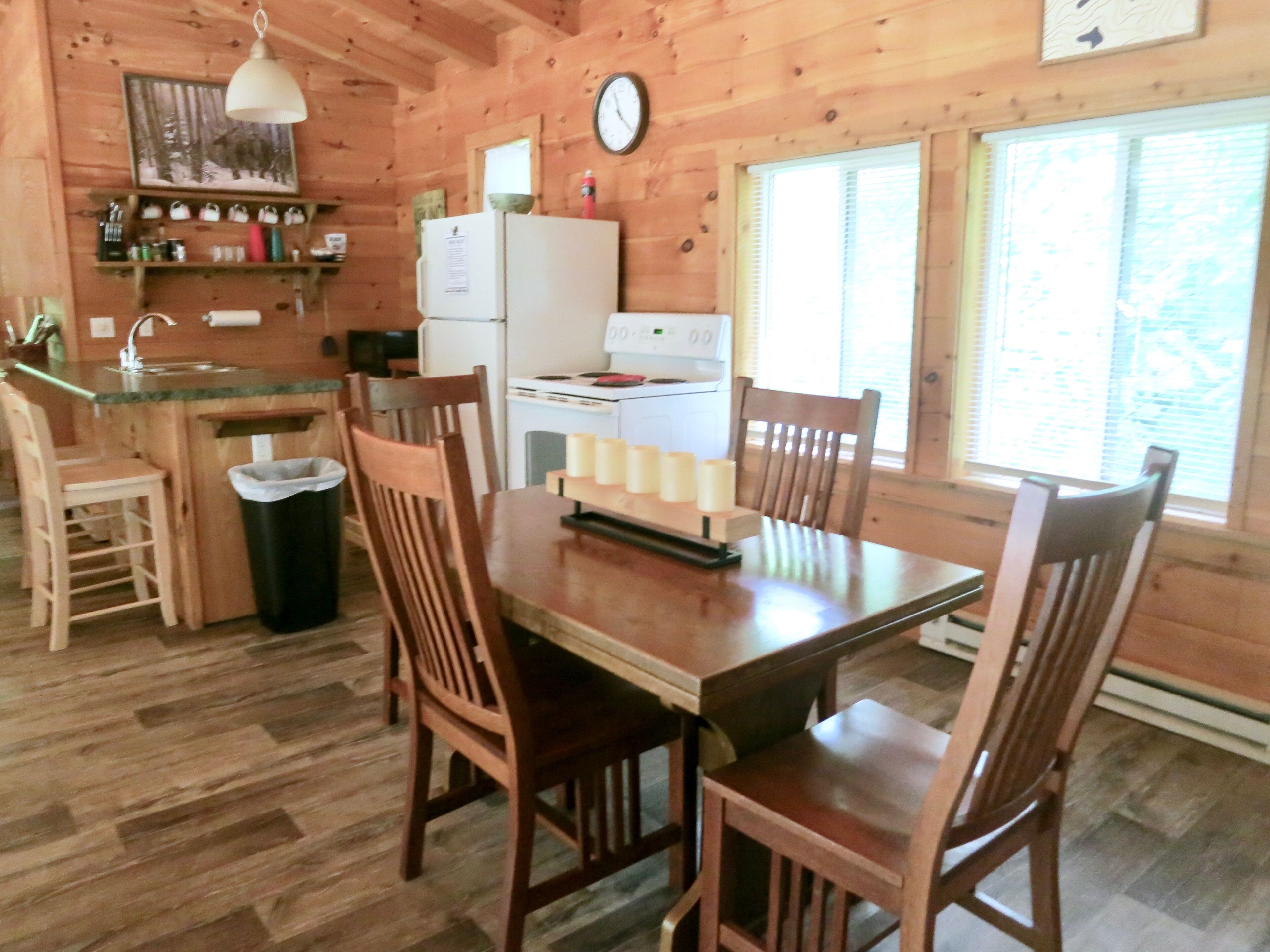 A wooden cabin interior with a kitchen, dining table, chairs, and a clock on the wall.