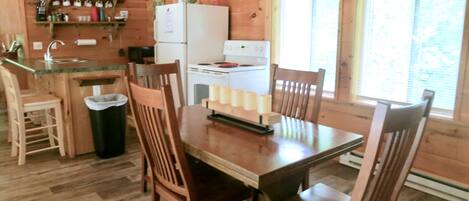 A wooden cabin interior with a kitchen, dining table, chairs, and a clock on the wall.
