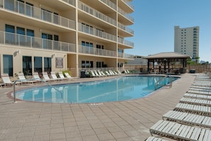 Une piscine dotée de chaises longues et d’un espace de repos couvert, le tout entouré d’un immeuble à plusieurs étages avec des balcons.