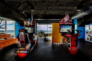 A room with a NASCAR racing simulator, a large TV screen, and a view of the ocean through large windows.