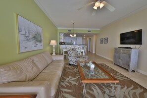 A living room with a sofa, a coffee table, and a television. There is a kitchen in the background with a dining table and chairs.
