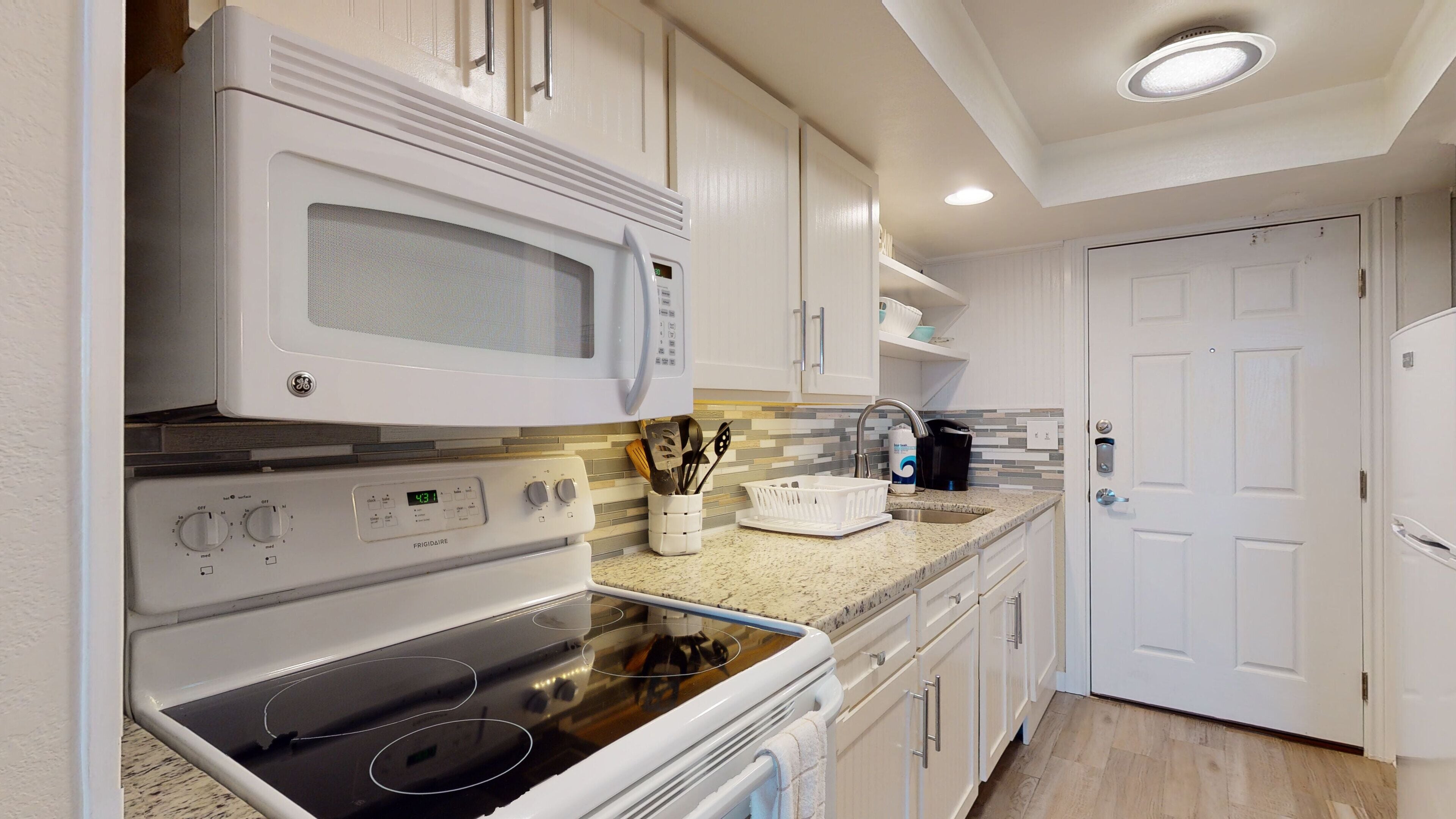 A compact kitchen with white appliances, including a microwave, oven, and dishwasher. There is a sink with a faucet, a countertop with a dish rack, and a door leading to another room.
