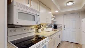 A compact kitchen with white appliances, including a microwave, oven, and dishwasher. There is a sink with a faucet, a countertop with a dish rack, and a door leading to another room. - Studio Condo in Town, Near Beach, Shared Pool and Hot Tub (Port Aransas)