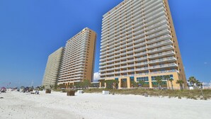 Two high-rise buildings with multiple balconies, a sandy beach in the foreground, and palm trees.