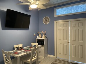 A room with a dining table, chairs, a TV mounted on the wall, a ceiling fan, a clock, a door, and a window with a view.