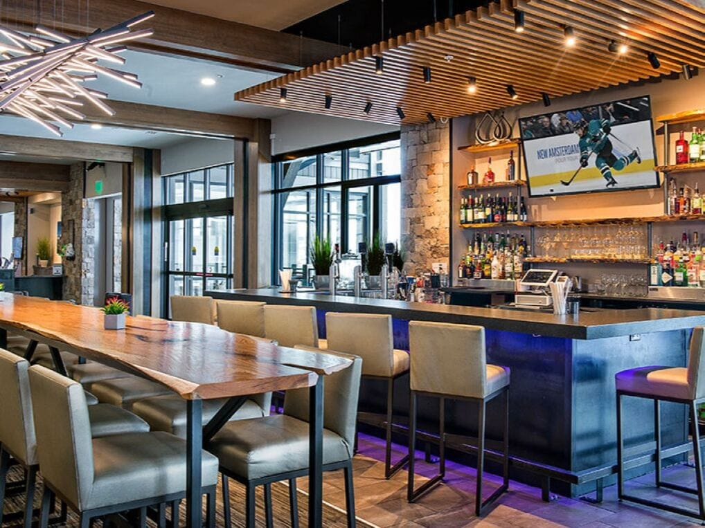 A bar area with a long wooden table and bar stools, a television screen displaying a hockey game, and a bar stocked with various bottles.