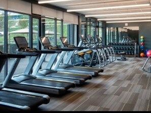 Een sportschool met loopbanden langs een muur met ramen, een houten vloer en fitnessballen in de hoek.
