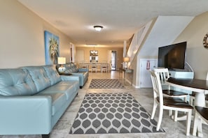 A living room with a blue sofa, a dining table with chairs, and a kitchen in the background.