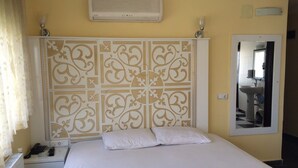 Habitación de hotel con cama, mesita de noche con lámpara, espejo y cabecera decorativa.