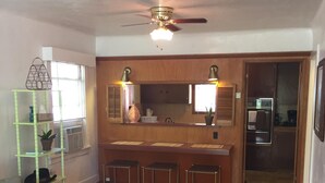 A living room with a wooden floor, a ceiling fan, a sofa, a coffee table, a patterned rug, a kitchen area with a refrigerator, and a wooden cabinet.