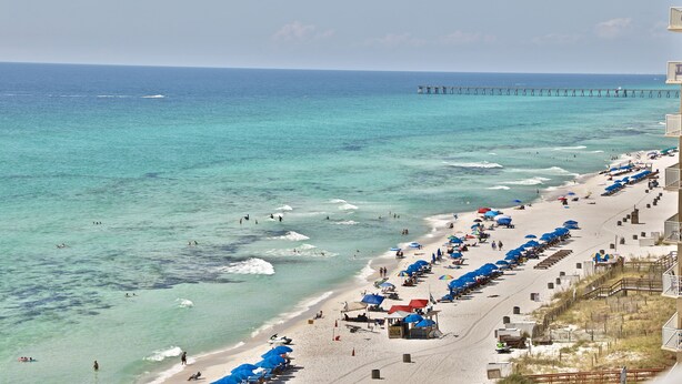 Ein Strand mit klarem türkisblauem Wasser, einem Pier in der Ferne und Reihen von Sonnenschirmen am Ufer.
