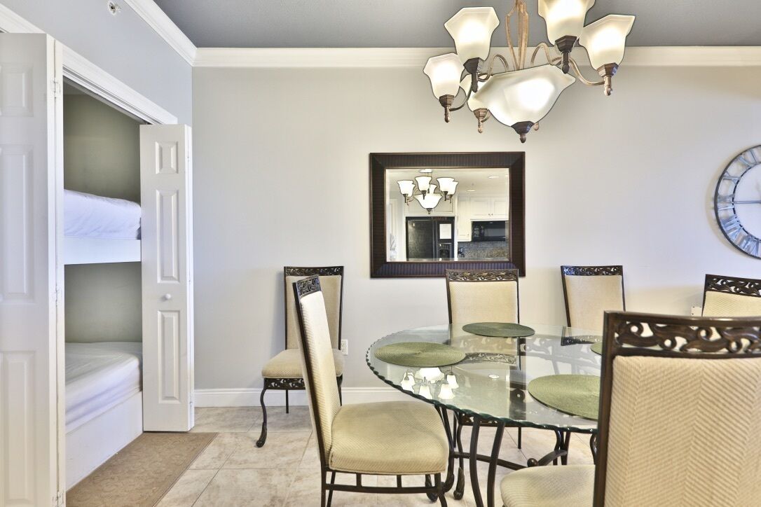 A dining area with a glass table, chairs with cushioned seats, a chandelier, and a wall clock.