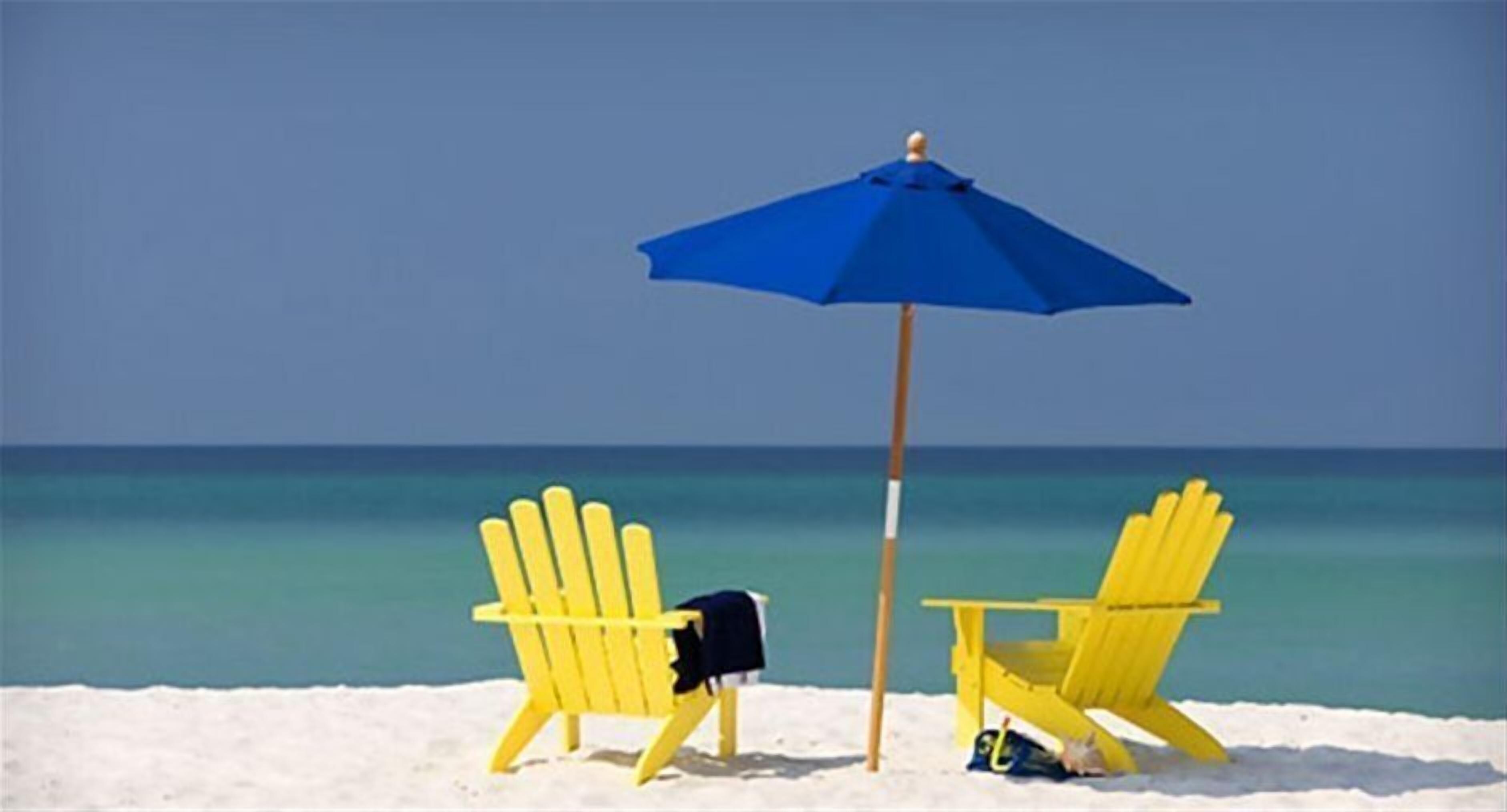 Two yellow Adirondack chairs on a sandy beach with a blue umbrella between them.