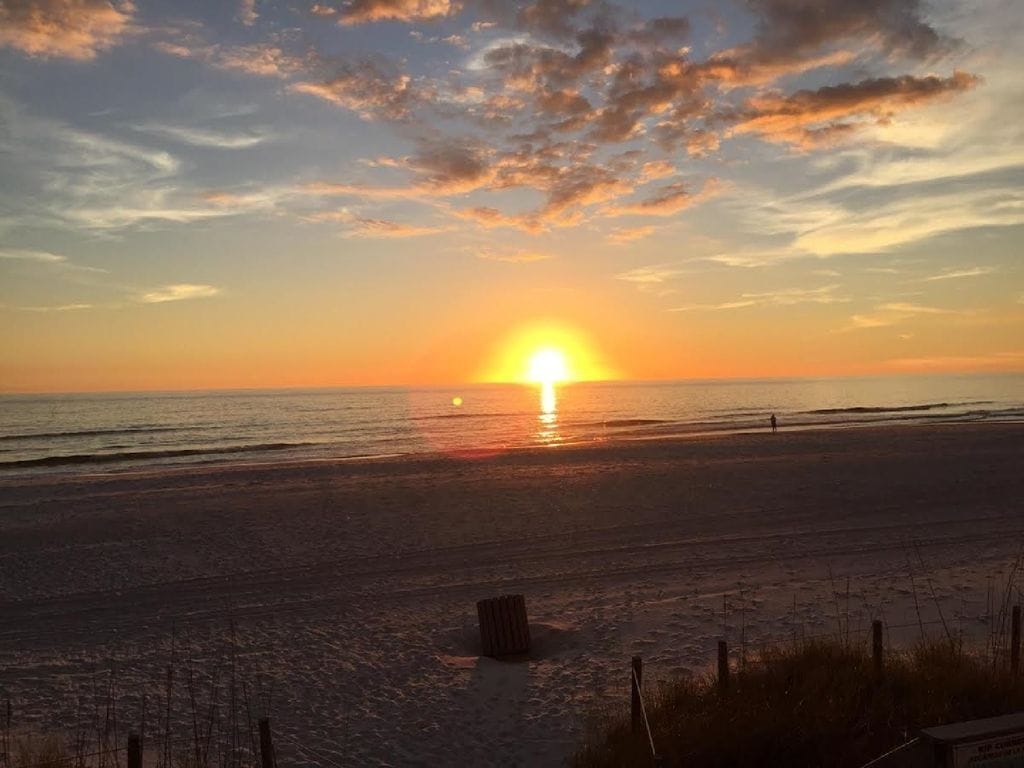 Une plage au coucher du soleil, avec le soleil bas à l’horizon, un ciel dégagé et une mer calme.