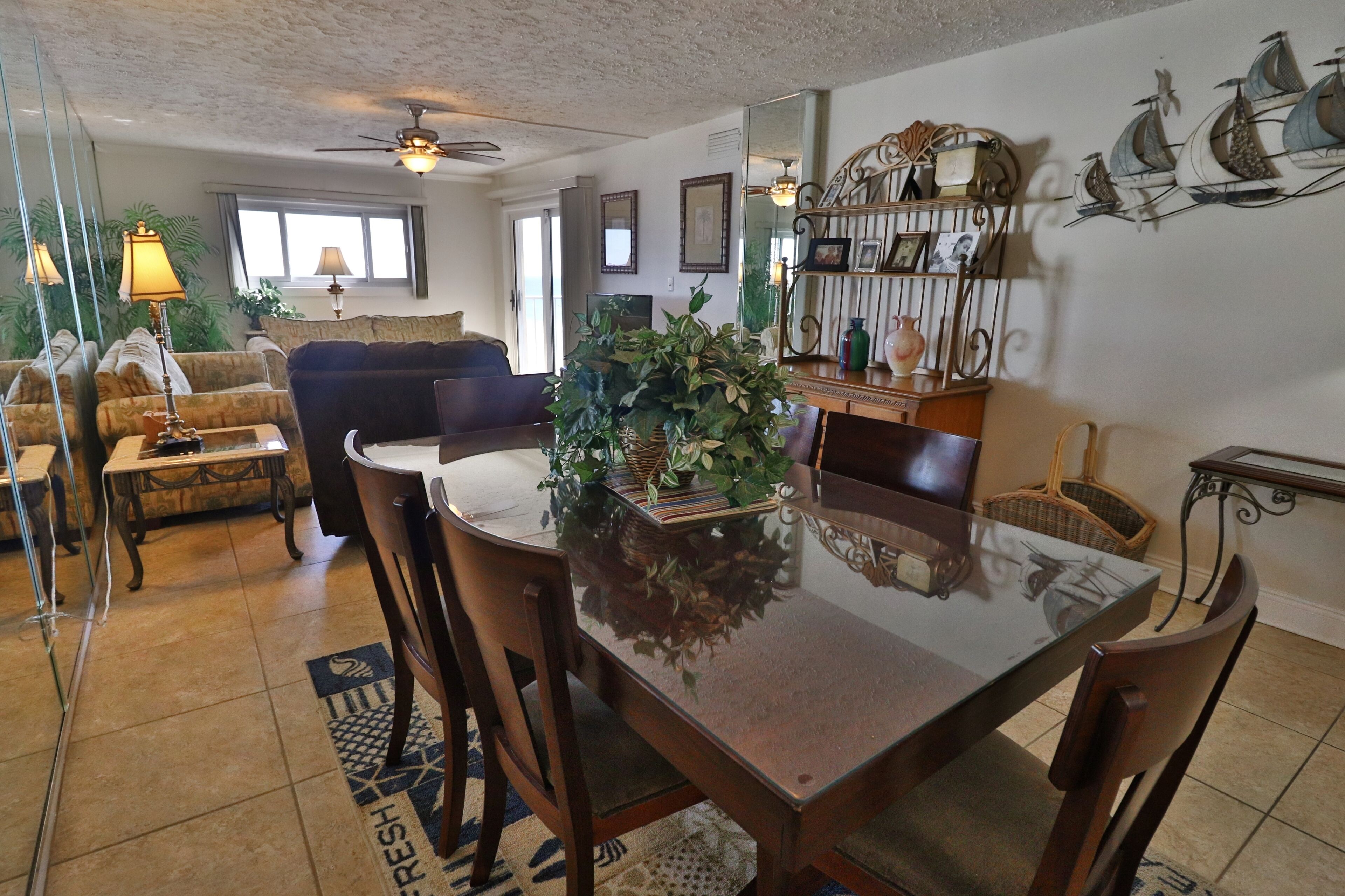 A dining area with a glass table, wooden chairs, and a central plant. There is a sofa and a window in the background.