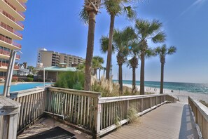 A wooden deck leading to a beach with palm trees, a pool, and a hotel building in the background.