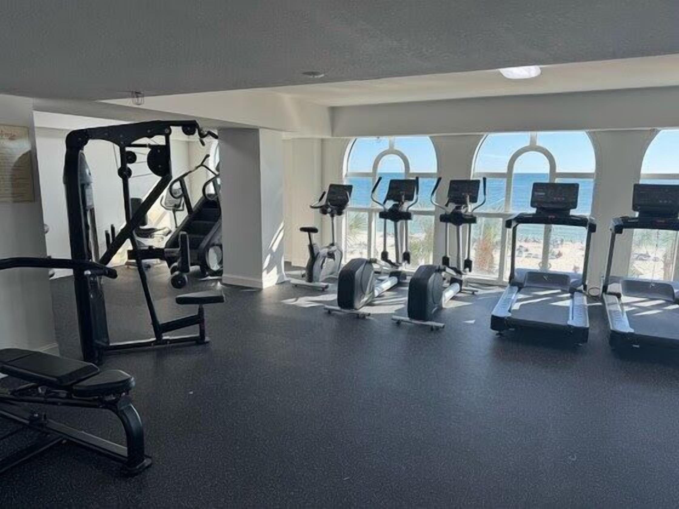 Un gimnasio con diversos equipos de ejercicio, amplias ventanas y vista al mar.
