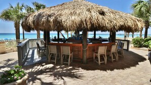 Zona de bar al aire libre con techo de paja, sillas y mesa de madera, con vistas a una playa con palmeras y un cielo azul despejado.