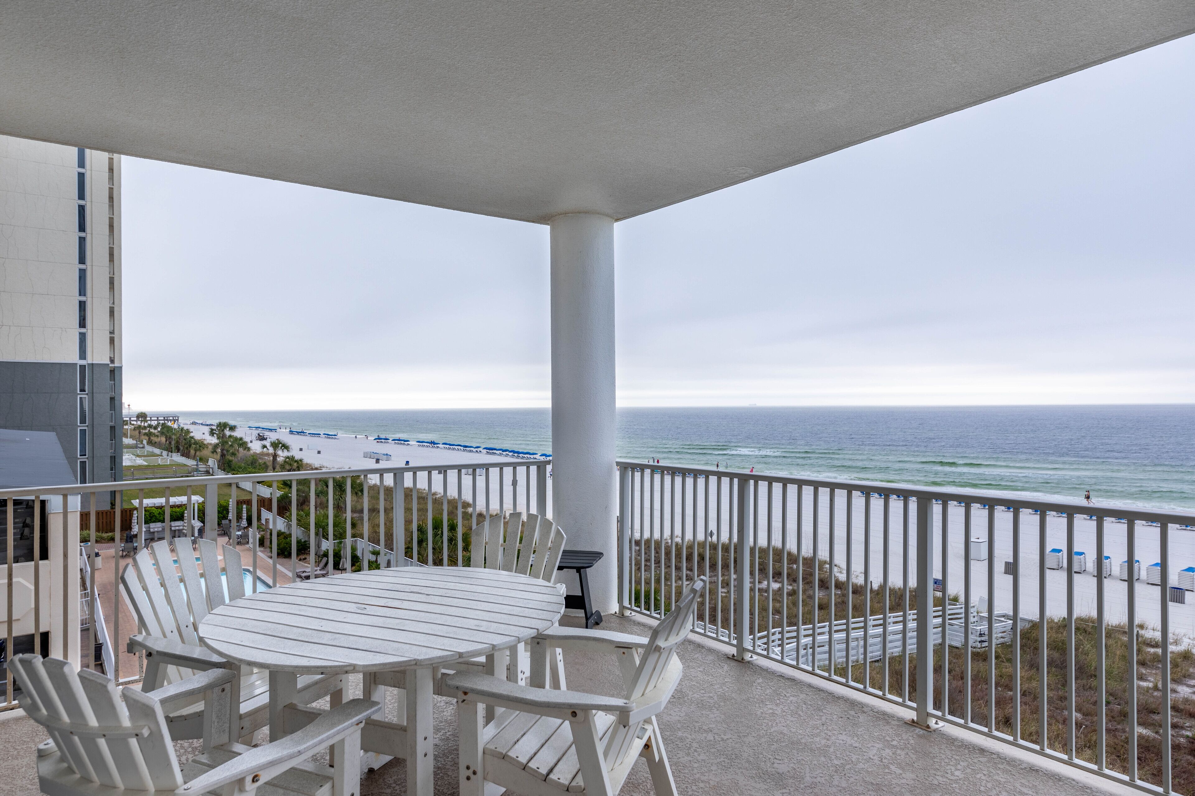 Un balcon avec une table et des chaises blanches, donnant sur une plage, avec vue sur l’océan et une jetée au loin.